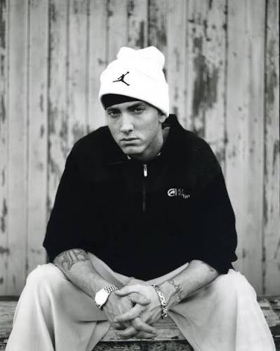 A rapper in a white beanie seated against a weathered wall